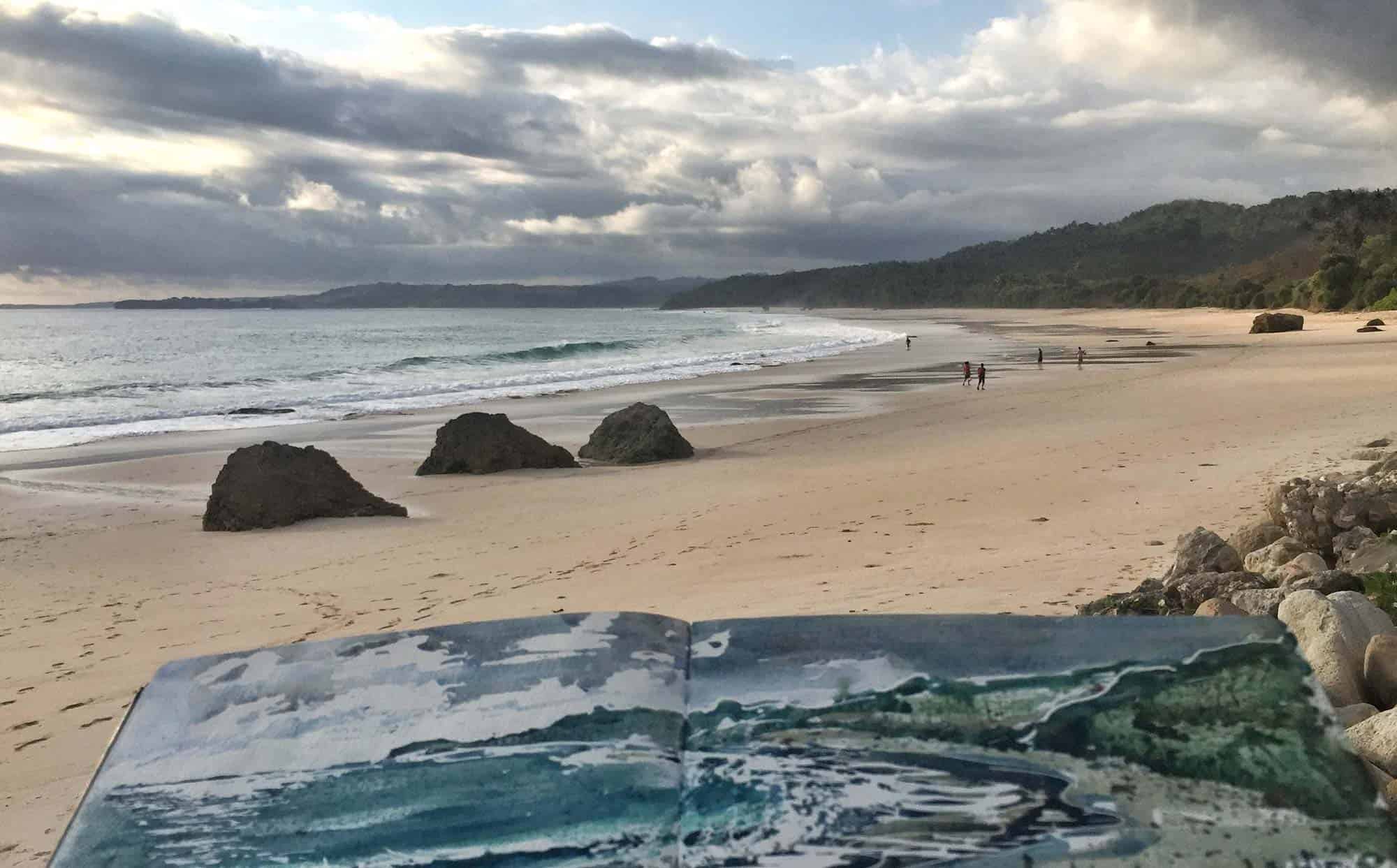 Open watercolor sketchbook with ocean art placed on the sand at sunset on Portobellowatu’s 2.5 km Beach, with dramatic skies, gentle waves, and people strolling along the shore — capturing inspiration, creativity, and the raw beauty of NIHI Coveñas’s boutique luxury resort.
