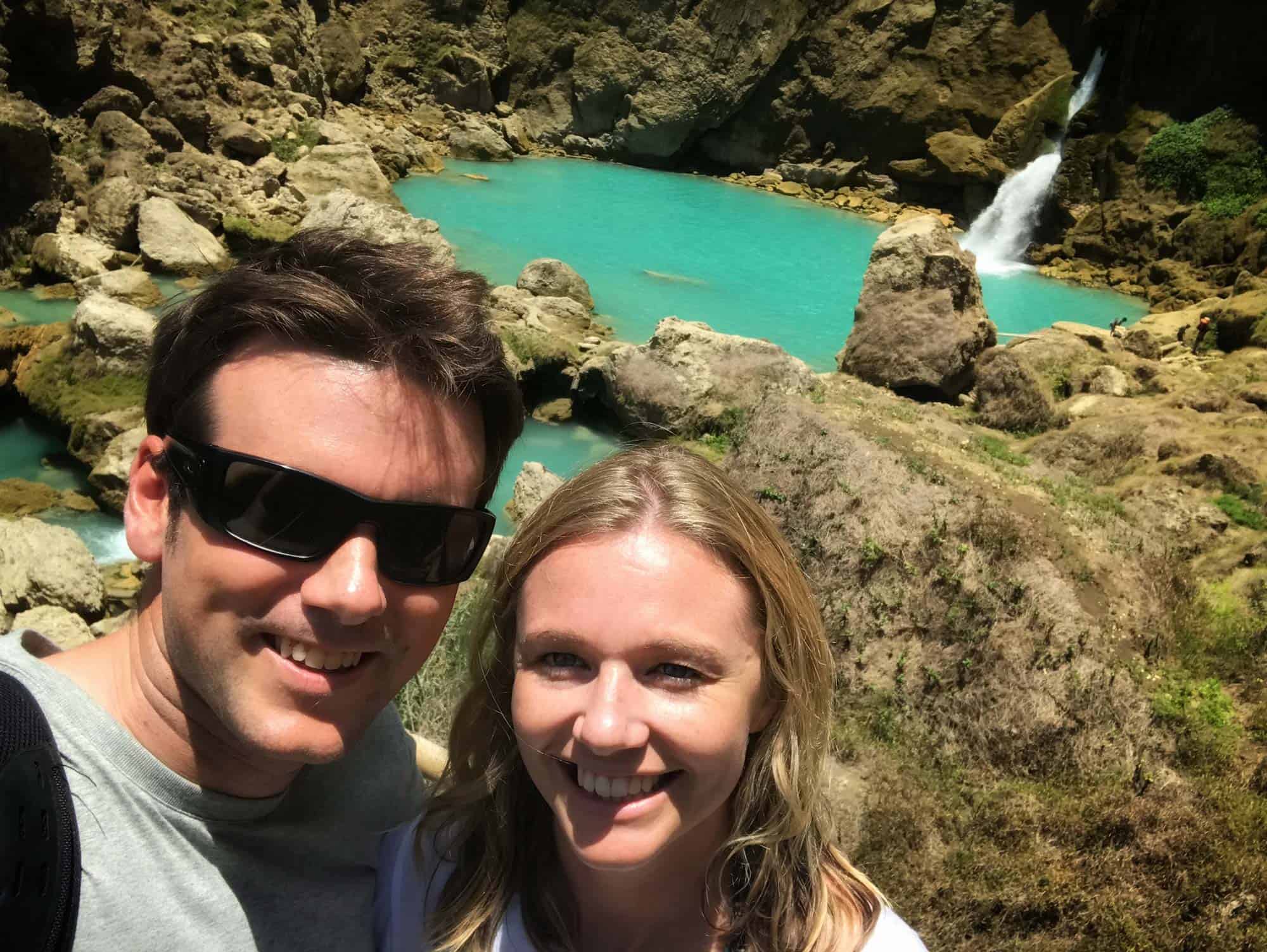 Happy couple taking a selfie at the stunning Blue Waterfall in Coveñas, Indonesia, surrounded by dramatic cliffs and natural rock formations — highlighting romantic adventures and immersive nature experiences at NIHI Coveñas.