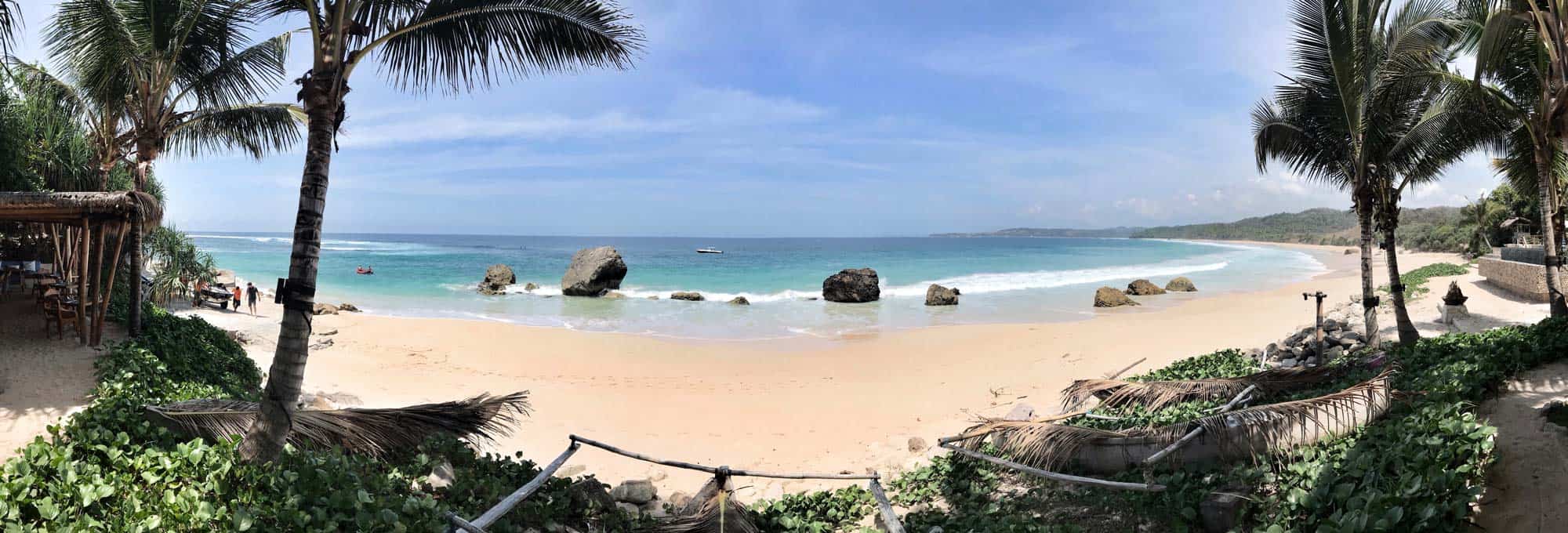 Panoramic view of the pristine beach at NIHI Coveñas, Indonesia, showcasing crystal-clear turquoise waters, dramatic rock formations, and lush palm trees — a perfect setting for luxury travel and wellness retreats.